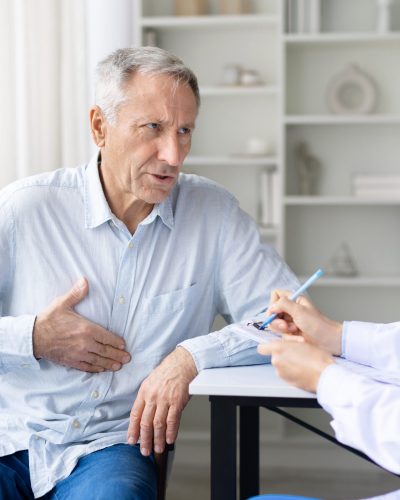 Senior male patient holding chest while talking to female doctor in white coat during medical consultation in clinic, healthcare and diagnosis concept